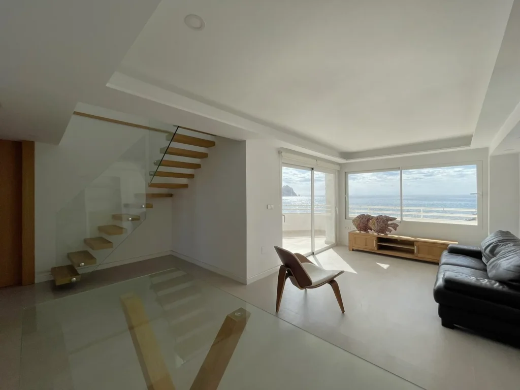 Salón del apartamento reformado: escalera volada de peldaños de madera con barandilla de vidrio y ventanales abiertos al mar
